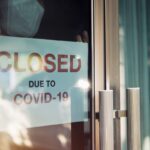 Business office or store shop is closed, bankrupt business due to the effect of novel Coronavirus (COVID-19) pandemic. Unidentified person wearing mask hanging closed sign in background on front door.