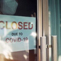 Business office or store shop is closed, bankrupt business due to the effect of novel Coronavirus (COVID-19) pandemic. Unidentified person wearing mask hanging closed sign in background on front door.