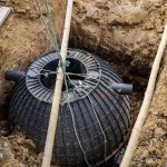 A large black underground tank, partially buried in a muddy excavation site. Two pipes extend from it, with wooden rods and ropes on top.